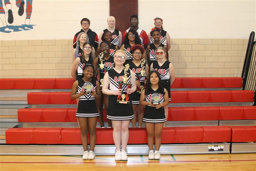 A group of cheerleaders in uniforms holding trophies on red bleachers in a gym.